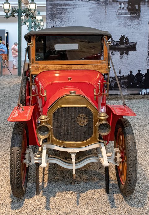 Front of a red two-seater with a black hood, brass radiator and two brass lamps on gravel. Opens a larger view