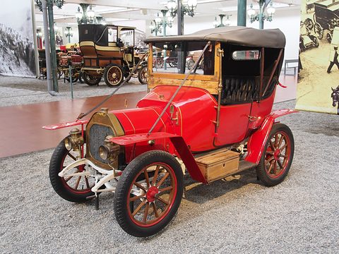 Red two-seater with a black hood, brass radiator and lamps and red wheels in a museum hall. Opens a larger view