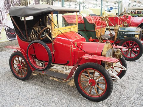 Red two-seater with a black hood, spare tyre and red wooden wheels on pale pebbles. Opens a larger view