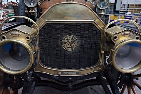 Close view of a brass radiator with a script badge flanked by two big acetylene headlamps. Opens a larger view
