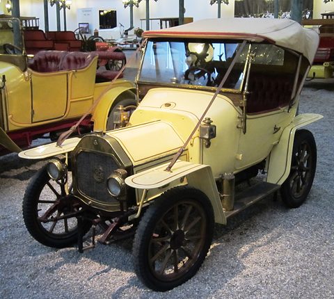 Cream two-seater with a beige hood, brass lamps and wooden wheels on gravel among other veteran cars. Opens a larger view