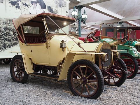 Cream two-seater with a folded hood frame, wooden wheels and brass lamps on pebbles. Opens a larger view