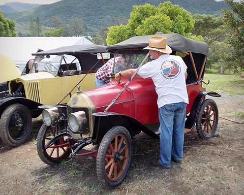 Red two-seater with wooden wheels on grass, a man in a white hat leaning over its bonnet. Opens a larger view