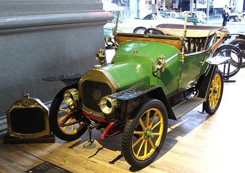 Green two-seater with yellow wheels and brass lamps on a wooden floor, a loose brass radiator beside it. Opens a larger view