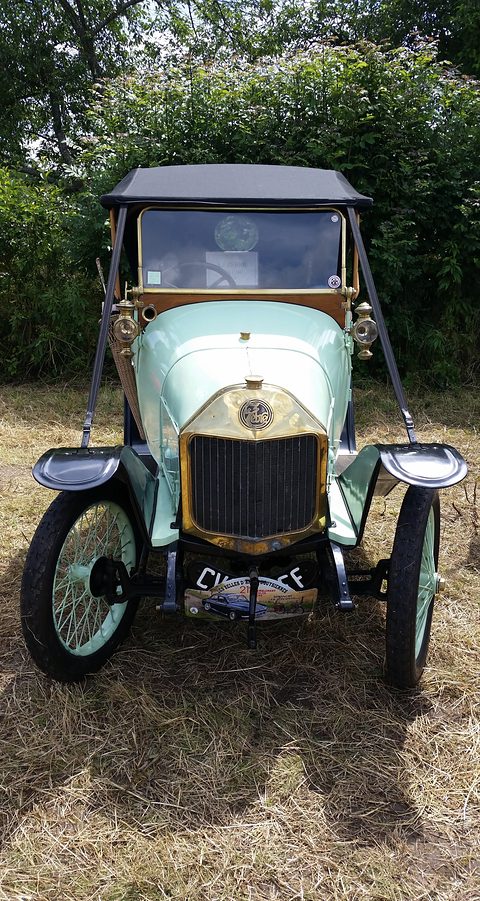 Front of a pale green two-seater with a black hood and brass radiator on cut grass. Opens a larger view
