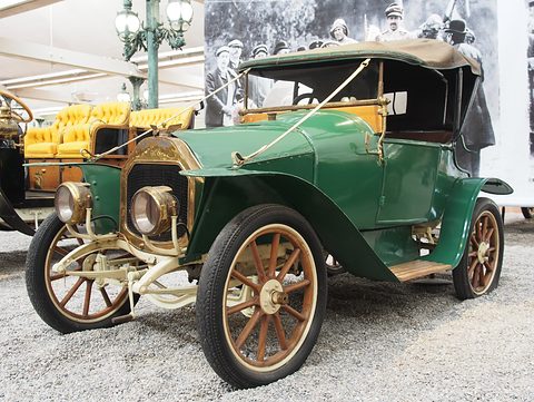 Green four-cylinder two-seater with a tan hood, brass lamps and wooden wheels on pebbles. Opens a larger view