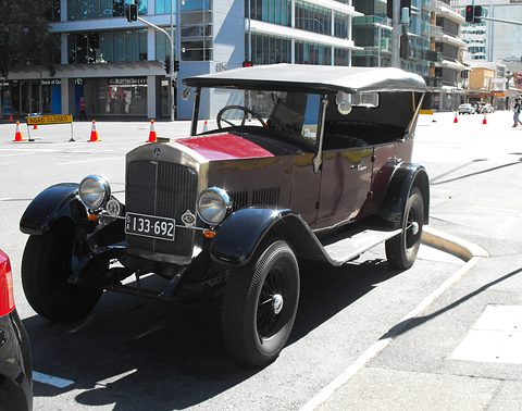 Black and maroon four-door open tourer with a cream hood parked on a sunny city street. Opens a larger view
