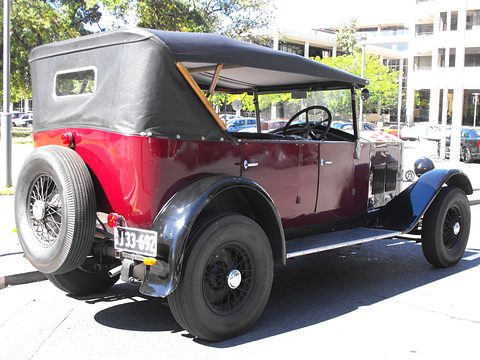 Rear of a maroon and black tourer with its hood up and a spare wheel on the back, in sunshine. Opens a larger view