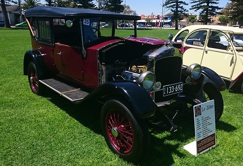 Maroon and black tourer with a black hood, bonnet side open, on a lawn beside a display board. Opens a larger view