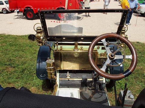 Looking over a wooden steering wheel and brass fittings to a folded windscreen and a car park. Opens a larger view