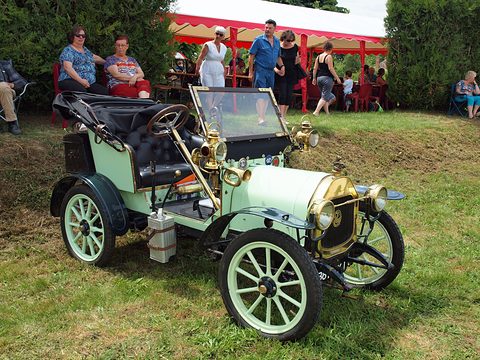 Pale green two-seater with brass lamps and pale wheels on grass, spectators under a red marquee. Opens a larger view