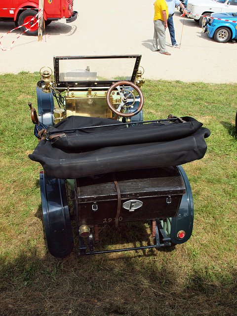 Rear of a small veteran car with a folded black hood and a brown trunk, on grass. Opens a larger view
