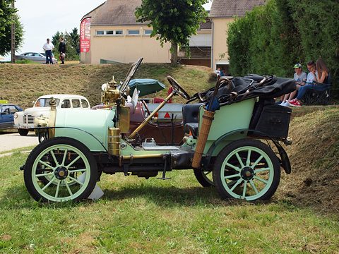 Side view of a pale green two-seater with brass trim and pale wheels on a grassy slope. Opens a larger view