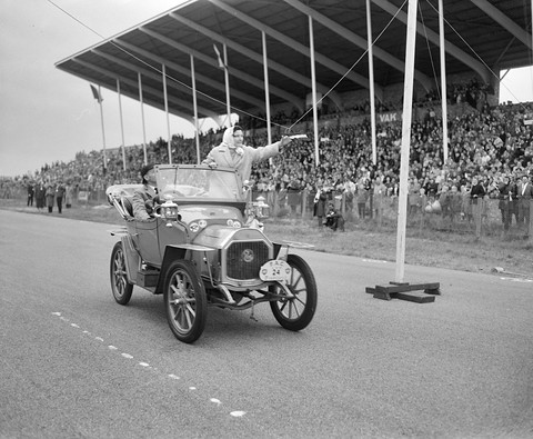 A small veteran car with a passenger standing to wave, passing a crowded grandstand on a track.