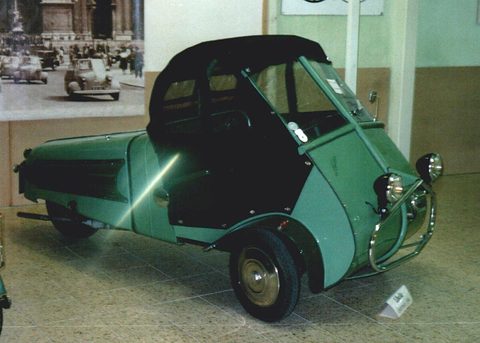 Green three-wheeler with a black fabric hood raised, a period street photograph on the wall behind. Opens a larger view