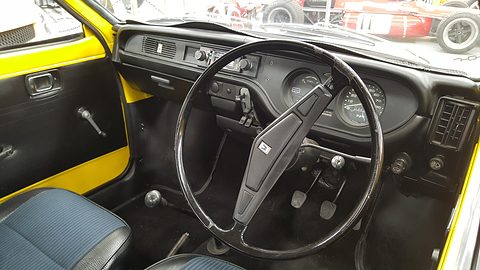 Black dashboard with a large two-spoke steering wheel, round dials and air vents, a yellow door panel and blue seat at the side. Opens a larger view