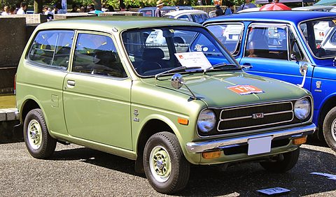 Green small three-door estate with round headlamps and a chrome grille, parked on a gravel field next to a blue car. Opens a larger view