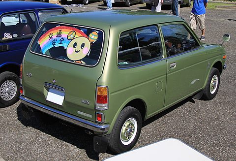 Rear quarter of a green small estate with a road-safety poster in its rear window, parked on gravel among other cars. Opens a larger view