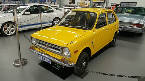 Yellow small two-door saloon with round headlamps, behind stanchions and a chain on a museum floor, with other classic cars around. Opens a larger view