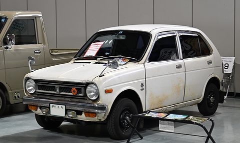 Weathered white small two-door saloon with round headlamps and a dark grille, on an indoor floor beside a pick-up truck. Opens a larger view