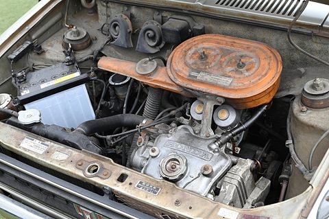 Open bonnet over a small engine with a large round orange air cleaner, a battery and hoses, the bodywork weathered. Opens a larger view