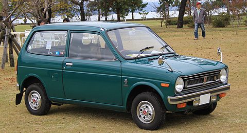 Dark green small three-door van with round headlamps and a chrome grille, parked on a dry lawn, a man walking behind. Opens a larger view