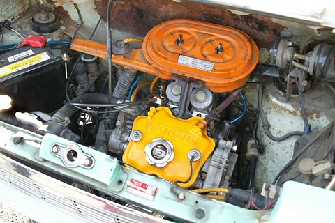 Engine bay with an orange round air cleaner and a yellow cam cover, blue wiring and a battery, the paint rusting at the edges. Opens a larger view