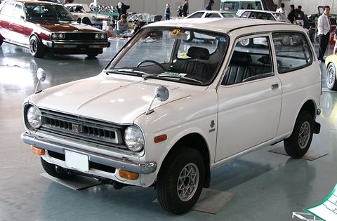 White small two-door saloon with round headlamps, a slatted grille and silver bumpers, on a polished floor among classic cars. Opens a larger view