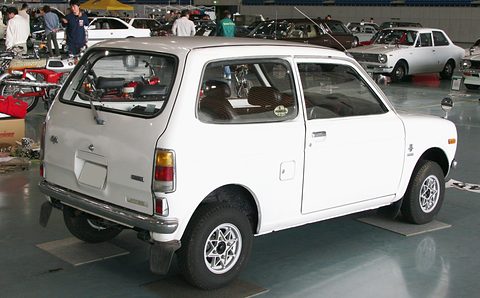 Rear quarter of a white small two-door saloon with alloy wheels and tall tail lamps, on a polished floor, visitors behind. Opens a larger view