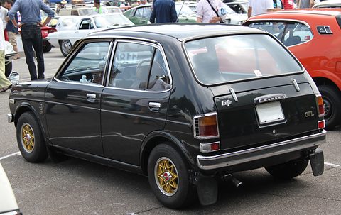 Rear quarter of a black small four-door saloon with gold wheels and a GFL badge, visitors and other cars behind. Opens a larger view
