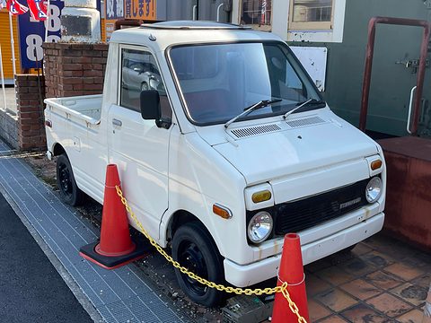 White small pick-up with a short nose, round headlamps and a tall cab, parked behind red traffic cones and a chain. Opens a larger view