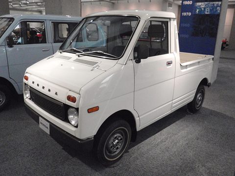 White small pick-up with a short nose, round headlamps and HONDA letters, on a museum floor beside a pale van.
