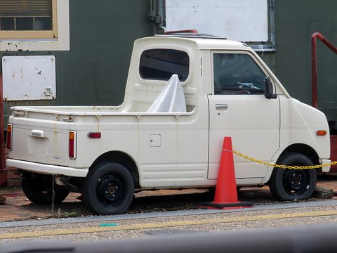 Rear quarter of a white small pick-up with a short load bed, parked behind a traffic cone and a chain in front of a green building. Opens a larger view