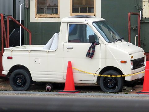 Side view of a white small pick-up with a tall cab and black wheels, behind red cones and a chain in front of a green wall. Opens a larger view