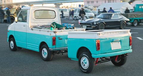 Turquoise and white small pick-up seen from behind, towing a small two-wheeled trailer in the same colours across a car park. Opens a larger view