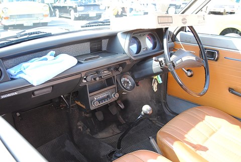 Small car cabin: black dashboard with round dials and a radio, thin steering wheel, tan door panels and seats, a cloth on the dash. Opens a larger view