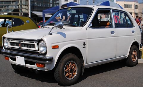 White small four-door saloon with a chrome grille, round headlamps and bronze wheels, in front of a green car and a blue tent. Opens a larger view