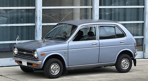 Pale blue-grey small four-door saloon in side-on view before a glass-fronted building. Opens a larger view