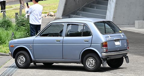 Rear quarter of a pale blue-grey small four-door saloon parked on concrete, a man with a phone by steps behind. Opens a larger view