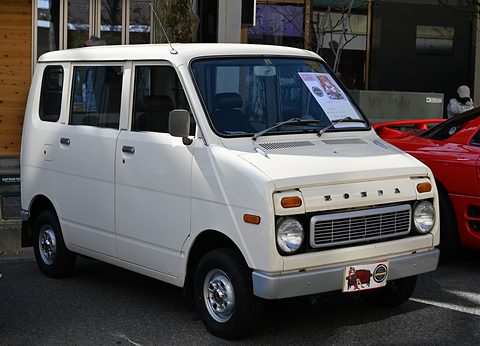 Tall white boxy small van with round headlamps and a slatted grille with Honda letters, a notice on the dashboard, a red car beside it. Opens a larger view