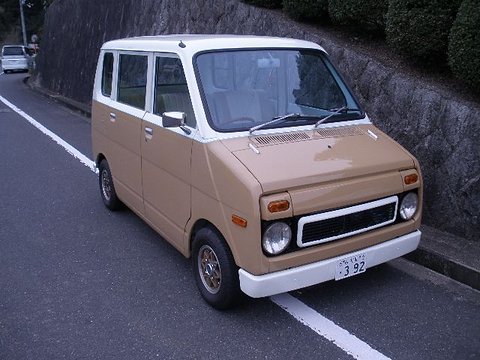 Beige tall small van with a white roof and bumper and a white grille surround, parked by a stone wall and hedges. Opens a larger view
