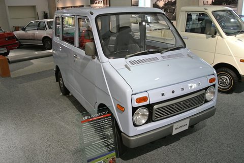 Pale grey tall small van with round headlamps and HONDA letters on its nose, beside an information panel on a museum floor.