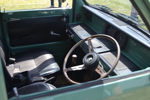 Green-painted cabin with a thin steering wheel, a long flat dashboard tray and black and white seats, seen through the open door. Opens a larger view
