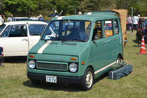 Pale green tall small van with a white stripe on its nose and a Japanese plate, parked on grass with a bag beside it. Opens a larger view