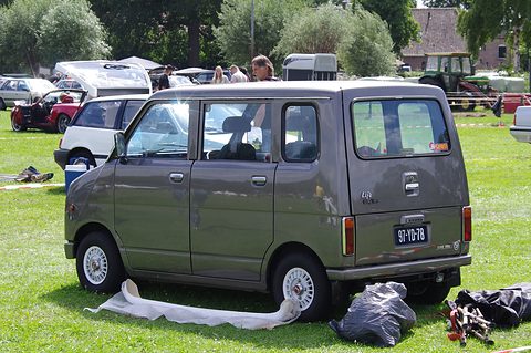 Rear quarter of a tall grey boxy small van with a large tailgate window, parked on grass with picnic bags beside a wheel. Opens a larger view