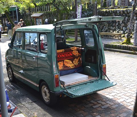 Rear of a green tall small van with its top-hinged tailgate and drop-down tail flap open, showing a Honda cushion inside.