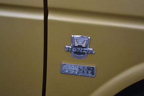 Chrome Honda H badge above a Super Deluxe script plate on mustard yellow paint, beside a door shut line. Opens a larger view