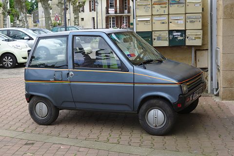 Small grey-blue hatchback with orange pinstripes parked on red block paving beside a wall of letterboxes. Opens a larger view