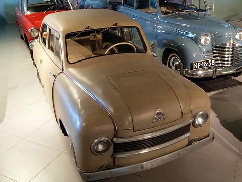 Beige small saloon seen from above the front, with slatted grille, triangle badge and pale steering wheel inside.