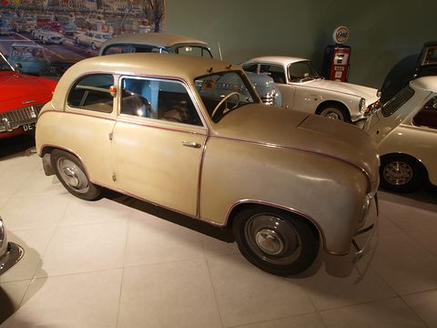 Beige small two-door saloon with red-piped seams on a pale tiled museum floor among other cars.